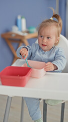 Fototapeta premium Toddler holds blue spoon to mouth while sitting in a highchair with pink and red bowls in building; curious play.