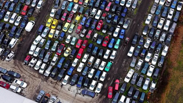 Overhead shot of scrapped vehicle lot. Tightly packed rows of damaged white, blue and red cars viewed from directly above a dirt yard.