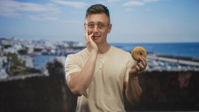 Man holding donut and touching cheek in a marina by the pier and sea, wearing glasses and a casual tshirt; hesitation hunger.