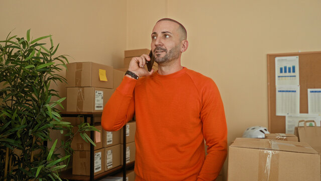 Man in orange sweater holds smartphone to ear with hand on cardboard box in parcel office building; small business determination.