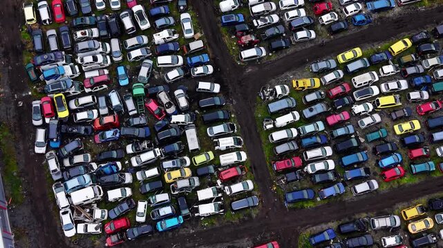 Top down view of parked scrap vehicles. Symmetrical rows of white, blue and yellow cars fill a gravel lot beside dry roadside grass.