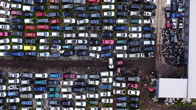 High angle aerial view of car dismantling site, Bulgaria. Curved rows of stripped vehicles and tire piles sit beside a warehouse rooftop below.