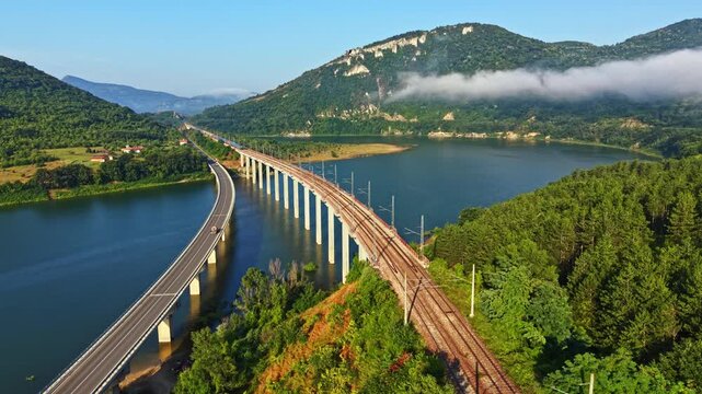 Elevated oblique view shows parallel bridges over calm river, with moving highway traffic on one side and empty railway tracks on other, framed by green hills, soft fog, and distant mountain peak
