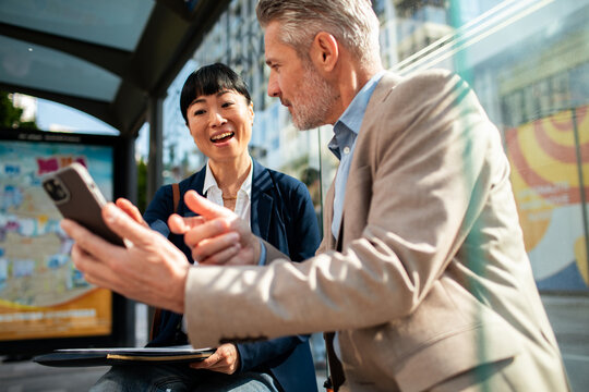 Two business colleagues discussing a smartphone at a city bus stop