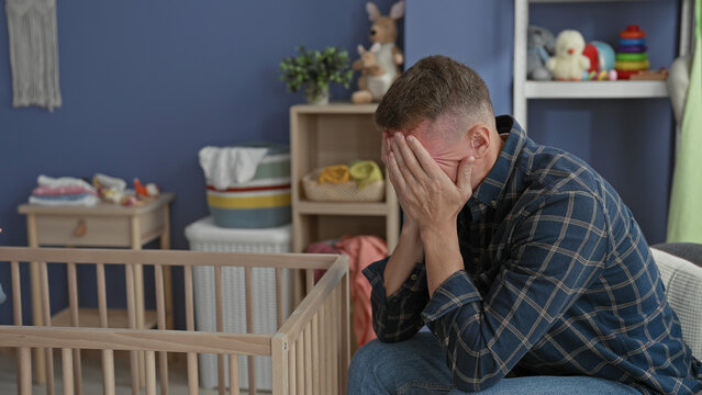 Man seated by crib in nursery with hands covering face and head bowed, toys on shelf and empty cradle visible in building; parenting stress despair.