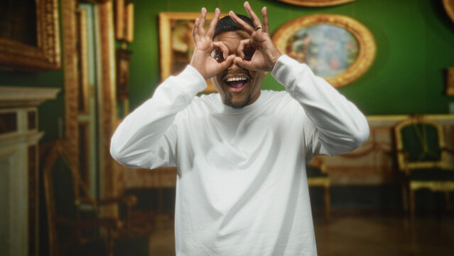 Man with beard wearing white shirt making ok sign to eye with hand while standing before ornate framed painting and antique chairs in building; playful joy.