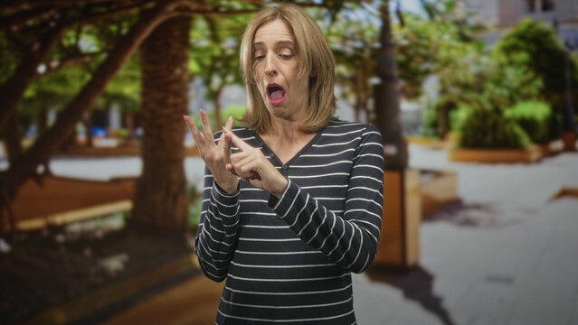 Woman inspects a fingernail on her hand in a busy street plaza, using the other hand to point and closely examine a possible hangnail or small injury; disgust concern.
