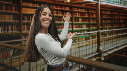 Woman with long hair smiling and gesturing with both hands toward wooden bookshelves in a library  joyful discovery. © Krakenimages.com
