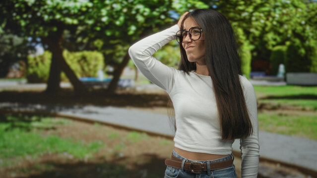 Woman with glasses hand behind head touching hair on street path in park; thoughtful doubt reflection.