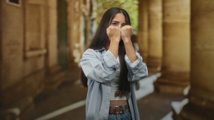 Fototapeta premium Woman with fists raised covering her face in a building colonnade walkway, hands tense and protective; anxiety resilience guarded determination.