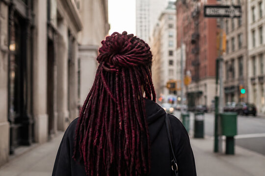 A person with long, red dreadlocks is standing on a city sidewalk in an urban environment.