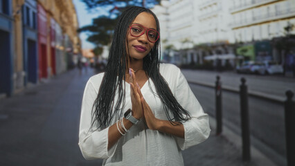 Fototapeta premium Woman with palms pressed together in prayer gesture on a street, wearing white blouse, red glasses, bracelets and smartwatch, braided hair and subtle smile; serenity.