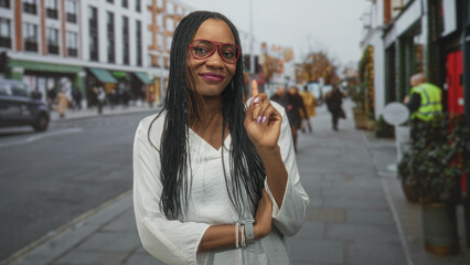 Fototapeta premium Black woman in white blouse wearing red glasses points finger on a street and shows bracelet and smartwatch; confidence warmth.
