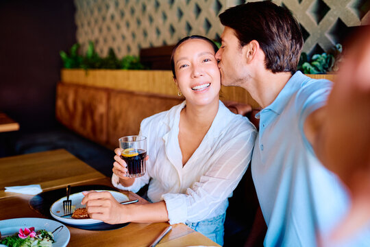 Cheerful couple sharing a meal, man kissing woman's cheek while she smiles at the camera holding a drink