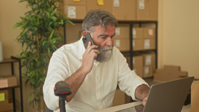 Man holding smartphone to ear and typing on laptop among packed boxes in a building; focused small business operations.
