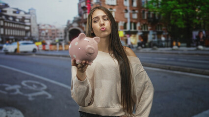 Obraz premium Young woman holds pink piggy bank in outstretched hand on a city street with bicycle lane markings; financial worry.