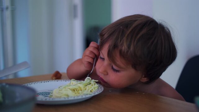 Child twirling spaghetti with fork at the kitchen table at home, focused self-feeding practice representing motor development, confidence and healthy independence