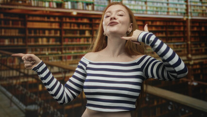 Redhead woman pointing finger left and making call me phone gesture at ear while wearing striped off shoulder crop top in a library building with bookshelves and railing  playful. © Krakenimages.com
