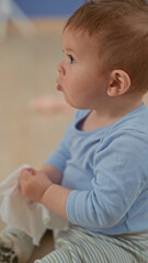 Fototapeta premium Hispanic toddler boy sits holding a soft cloth, fingers clasped and looking sideways while wearing a blue top in a building; curiosity warmth.