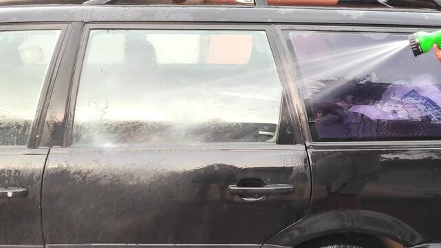 Person cleaning car window with hose spray, woman washing vehicle window with water nozzle outdoors, car maintenance.