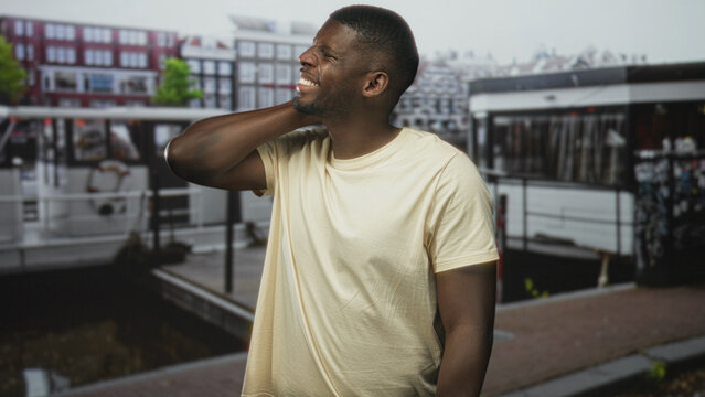 Man holding neck for backache and grimacing on street by a canal boat and dock, with nearby buildings and pavement visible; discomfort.