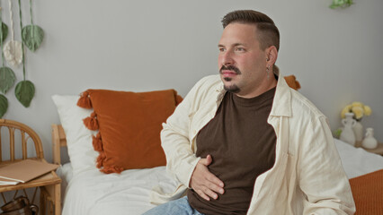 Fototapeta premium Man sitting on bed in studio with hand on stomach clutching abdomen and squinting in pain, suggesting stomachache; discomfort.