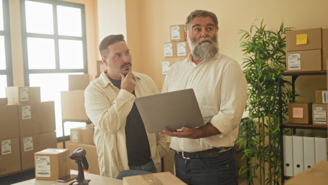 Two men review parcels on a laptop, younger man with hand on chin while older man holds laptop inside building; small business planning focused.