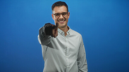 Naklejka premium Man with moustache in striped shirt smiles and points finger at camera, hand visible in studio backdrop with blue wall; playful charm.