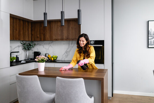 Smiling Caucasian American woman cleaning modern kitchen countertop with spray bottle and pink gloves