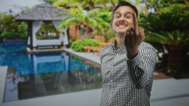 Young man with moustache points and shows middle finger toward camera by poolside building; defiance.