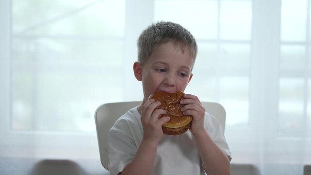 Happy kid eating burger at home. Close-up of kid enjoying food. Smiling boy loves his fast burger. Joyful child savors delicious burger. Kid eating tasty food. Kid delight in enjoying fast food