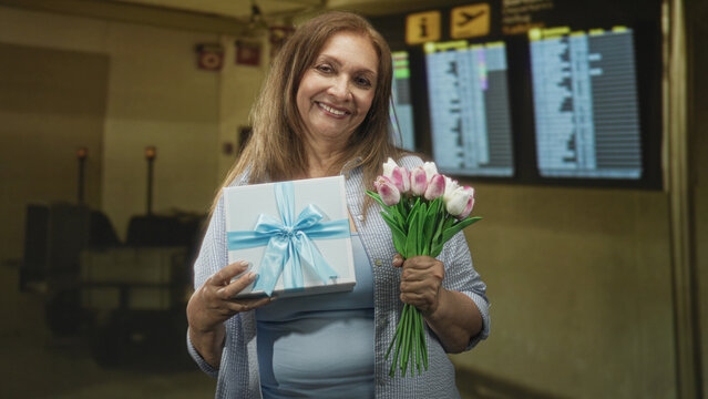 Senior hispanic woman holds blue ribbon gift box and pink tulip bouquet while smiling in an airport check in area by the departure display; joy reunion celebration.