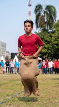 A young man in a red t-shirt participates in a traditional sack race competition during Indonesian Independence Day celebrations outdoors.