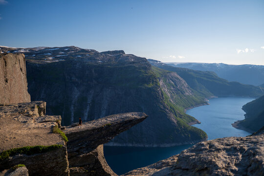 Aerial view of Trolltunga rock formation overlooking a blue fjord lake and steep mountain slopes in Norway under clear summer sky.