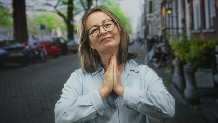 Fototapeta premium Woman senior with grey hair and glasses pressing palms together in prayer on a cobblestone street beside parked bikes and cars, wearing light blue shirt; gratitude hope.