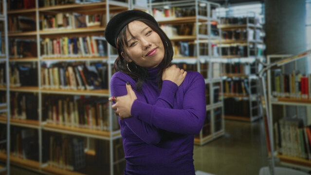Woman hugging her shoulders with hands, wearing beret and purple sweater in building library aisles, eyes closed and smiling; comfort.