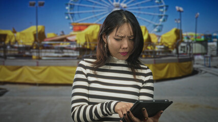 Fototapeta premium Young asian woman taps a tablet with hands at a street fairground by a ferris wheel; concentration curiosity engagement.
