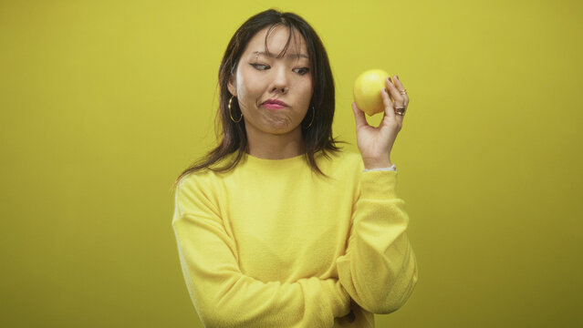 Woman holds lemon near face with raised hand, puckered lips and sideways glance, wearing yellow sweater and hoop earring against seamless yellow studio backdrop; skepticism.
