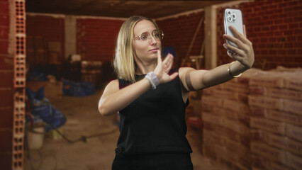 Obraz premium Woman taking selfie with smartphone, waves arm and pouts in building, wearing glasses, smartwatch and bracelets with exposed arm and stacked bricks nearby; playful moment.
