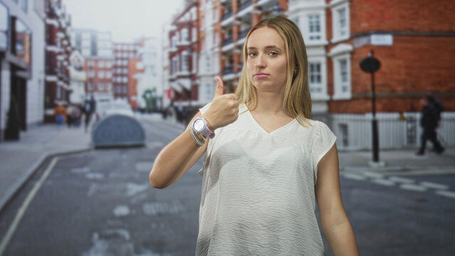 Woman giving a hesitant thumbs up while showing a wristwatch and white blouse on a city street lined with brick buildings and a lamppost.; ambivalence approval.