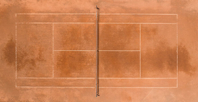 Top-down aerial view of a red clay tennis court with white marking lines and a net dividing the field in the center, showing a minimalist geometric sports layout. Zenithal shot of a empty court.