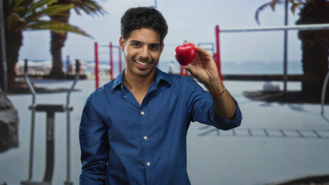 Man holding red heart stressball, smiling with eyes closed at a beach workout area with palm trees and pullup bars; love joy.
