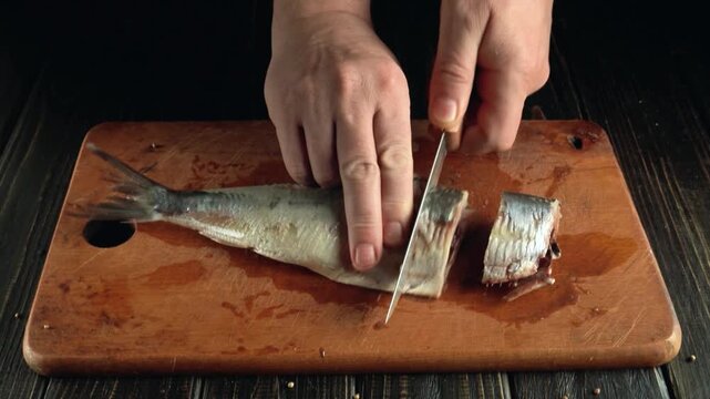 Hands of a person slicing a fish on a wooden cutting board, with visible fish parts and a clean kitchen surface, showcasing the progression of the cutting process in a culinary setting