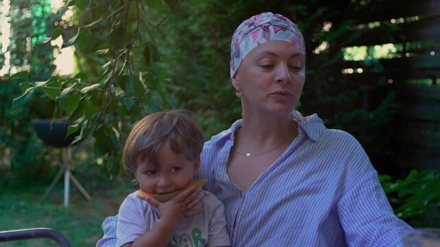 Mother with cancer holding toddler eating melon in a garden, nurturing outdoor family moment expressing resilience, connection and everyday joy through illness