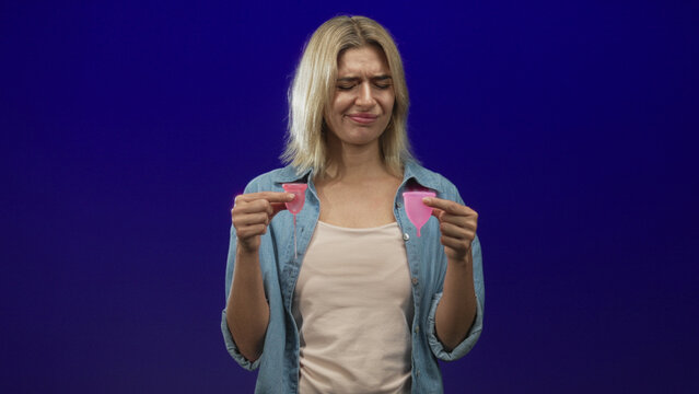 Woman holds two pink menstrual cups comparing sizes with a grimace and pinched fingers in a studio; period choice uncertainty.