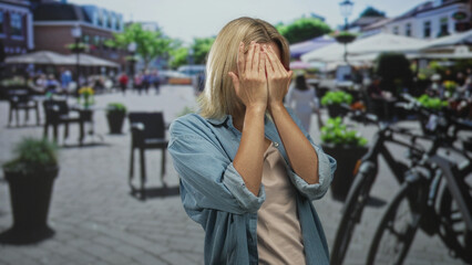 Fototapeta premium Young woman adjusts eyeglasses with both hands, smiling and showing bare forearms while standing in a busy street; casual city leisure joy.