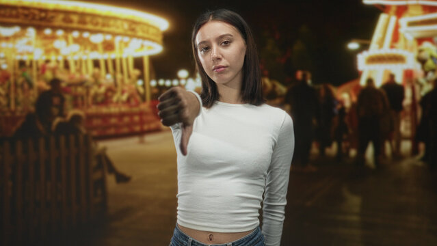 Young woman giving thumbsdown with extended thumb to camera at a nighttime carnival with carousel lights and blurred crowd; disapproval rejection.