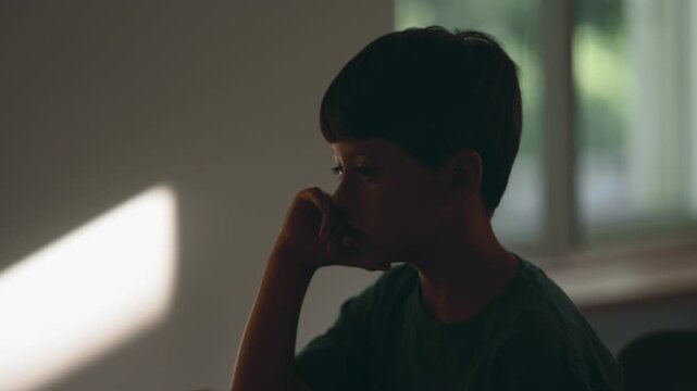 Sad depressed young boy sitting alone at home with downcast expression, lost in painful thoughts, portraying childhood depression, emotional isolation, anxiety, grief, hopelessness and mental health