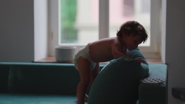 Toddler climbing cushion and falling onto sofa during indoor play, celebrating curiosity, confidence and joyful childhood exploration