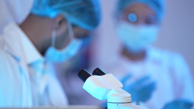 Gloved hands adjust a camera equipped microscope under moody blue light in a futuristic biotechnology laboratory as a masked colleague works nearby close up shot